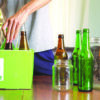 Person placing glass bottles into a recycling bin beside reusable glass containers.