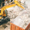 Excavator loading construction and demolition debris into a large dump truck at a dusty demolition site.
