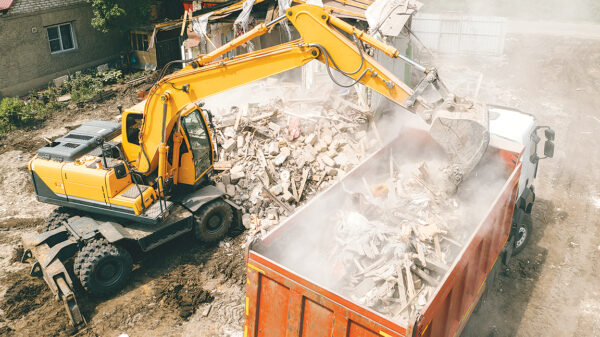 Excavator loading construction and demolition debris into a large dump truck at a dusty demolition site.
