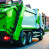 Green municipal garbage truck collecting waste along a residential street, illustrating waste collection route optimization.